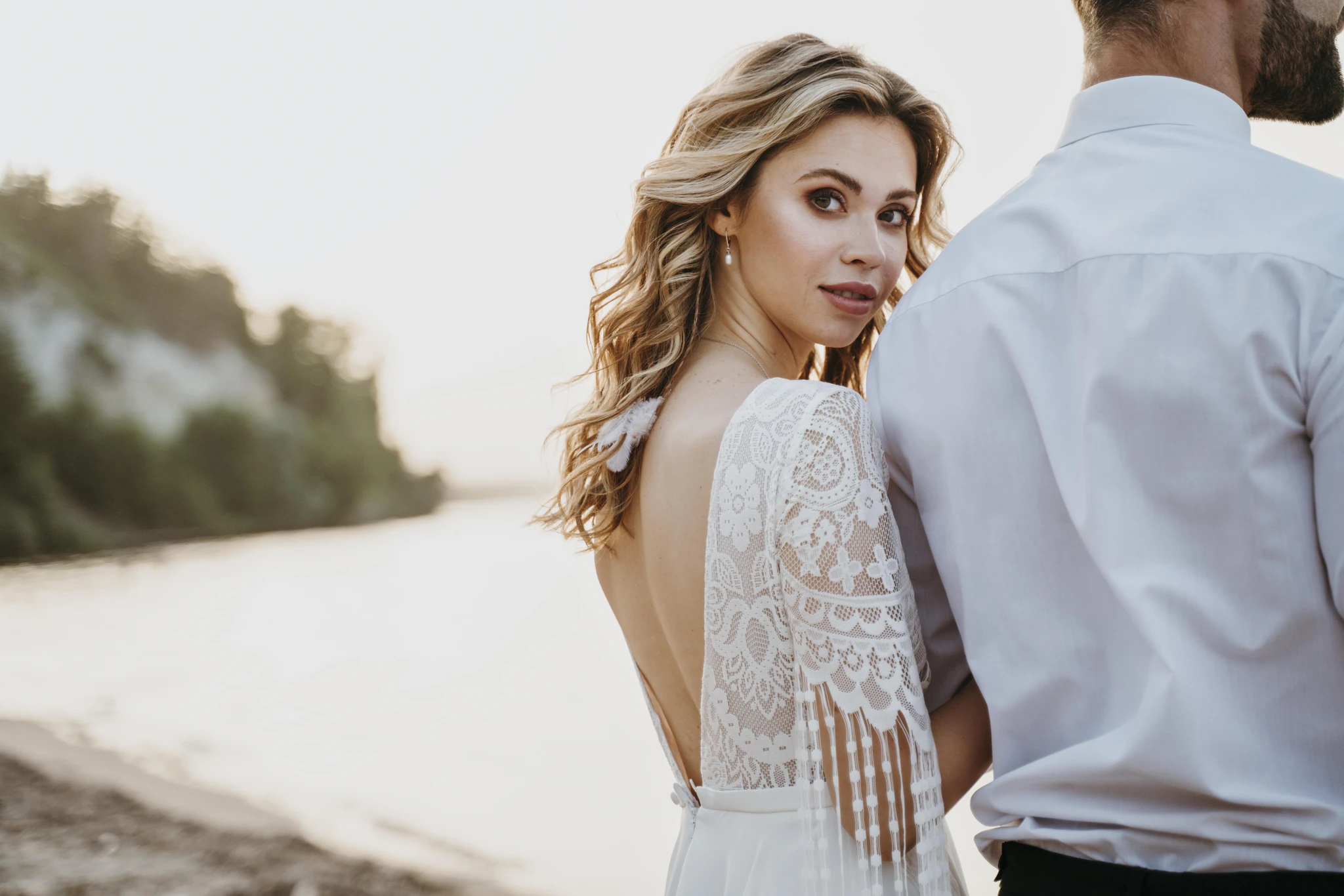 Bride in a lace dress looking back over her shoulder beside the groom on the shore