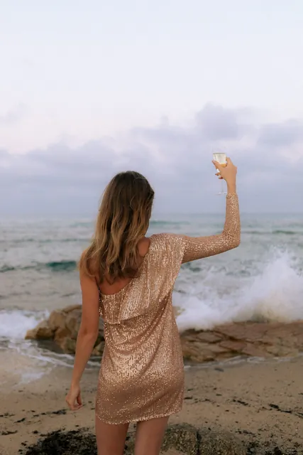 A woman in a gold sequin dress raising her glass towards the waves